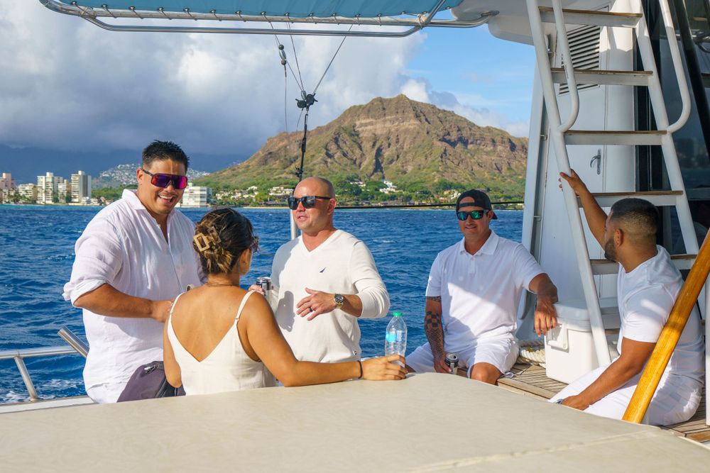 Group enjoying drinks and conversation on a yacht deck with ocean and mountains in background.