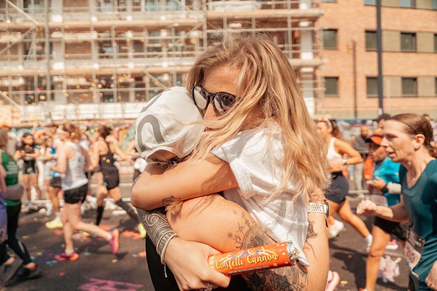 Two people embrace at an outdoor festival while a crowd gathers in the background near scaffolding and buildings.