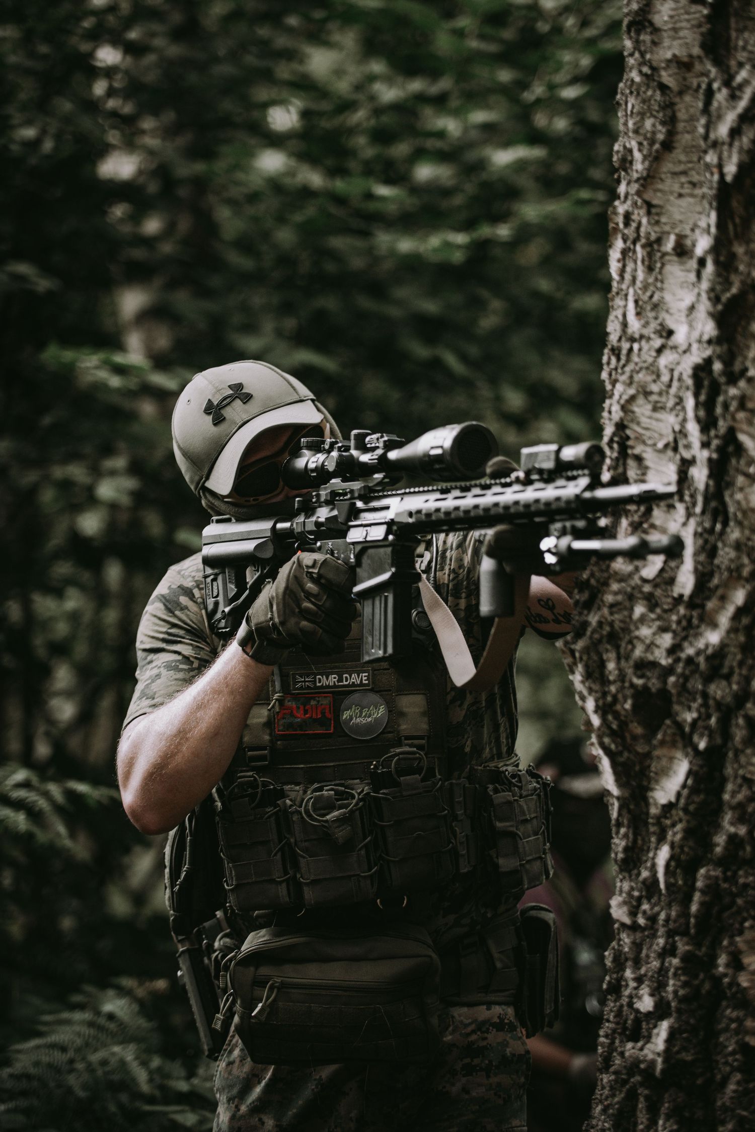 Tactical operator in dark gear aims rifle with scope from behind tree in moody forest setting.
