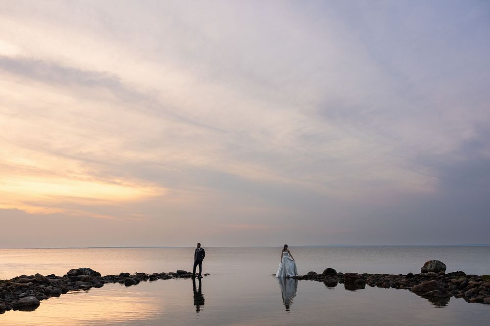 A wedding couple poses on rocky shores during a golden sunset over calm water. aKaiserPhoto.