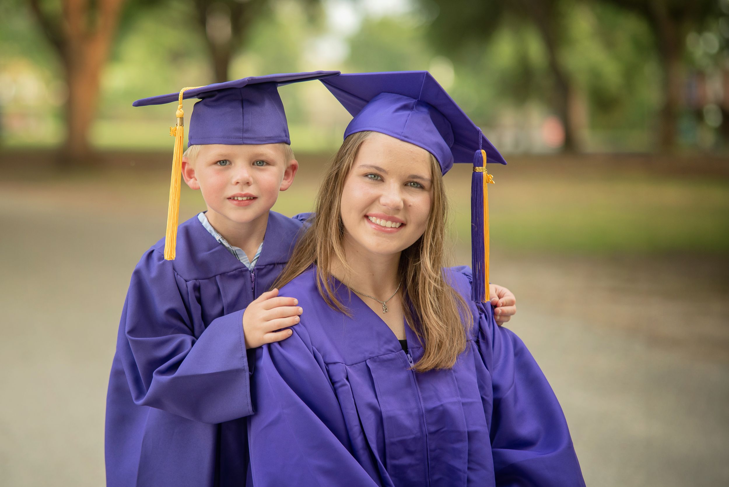 senior sister posing with kindergarten brother in cap and gown in park