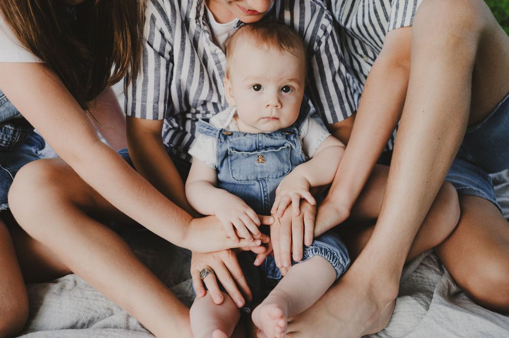 An infant in denim overalls sits between crossed legs on a comfortable surface.