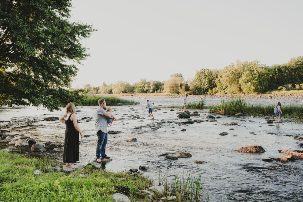 Two people stand by a rocky riverbank at sunset with trees in the background.
