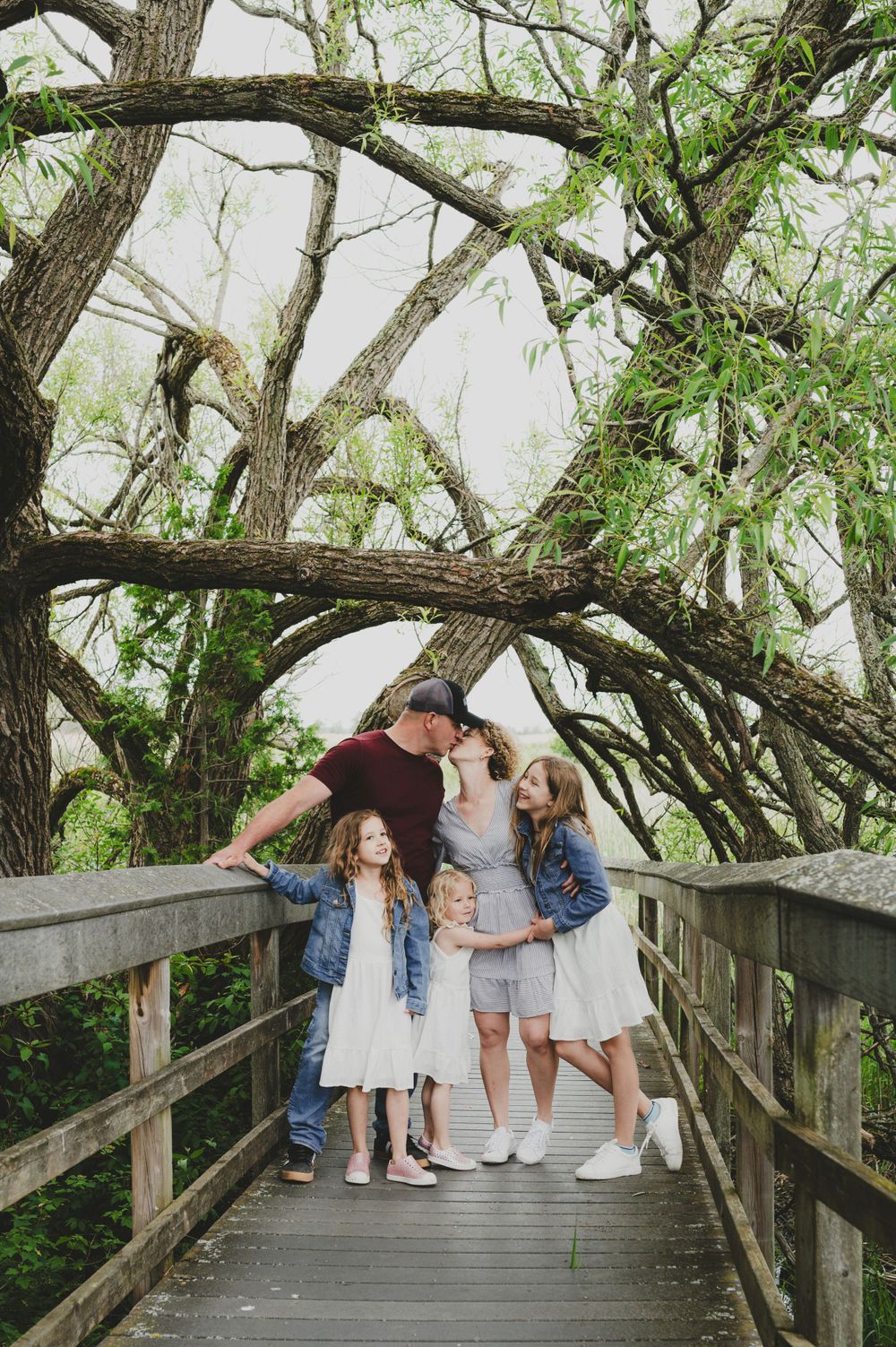 A family shares an intimate moment on a wooden bridge under arching tree branches.