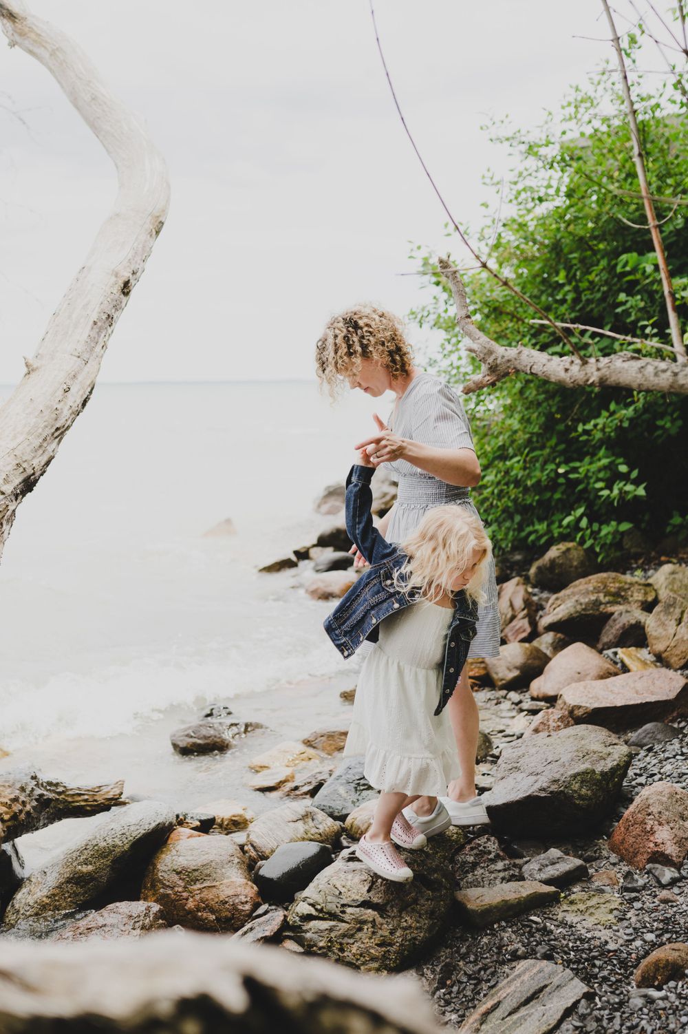 Young siblings explore a rocky shoreline beneath a white tree branch on a cloudy day.