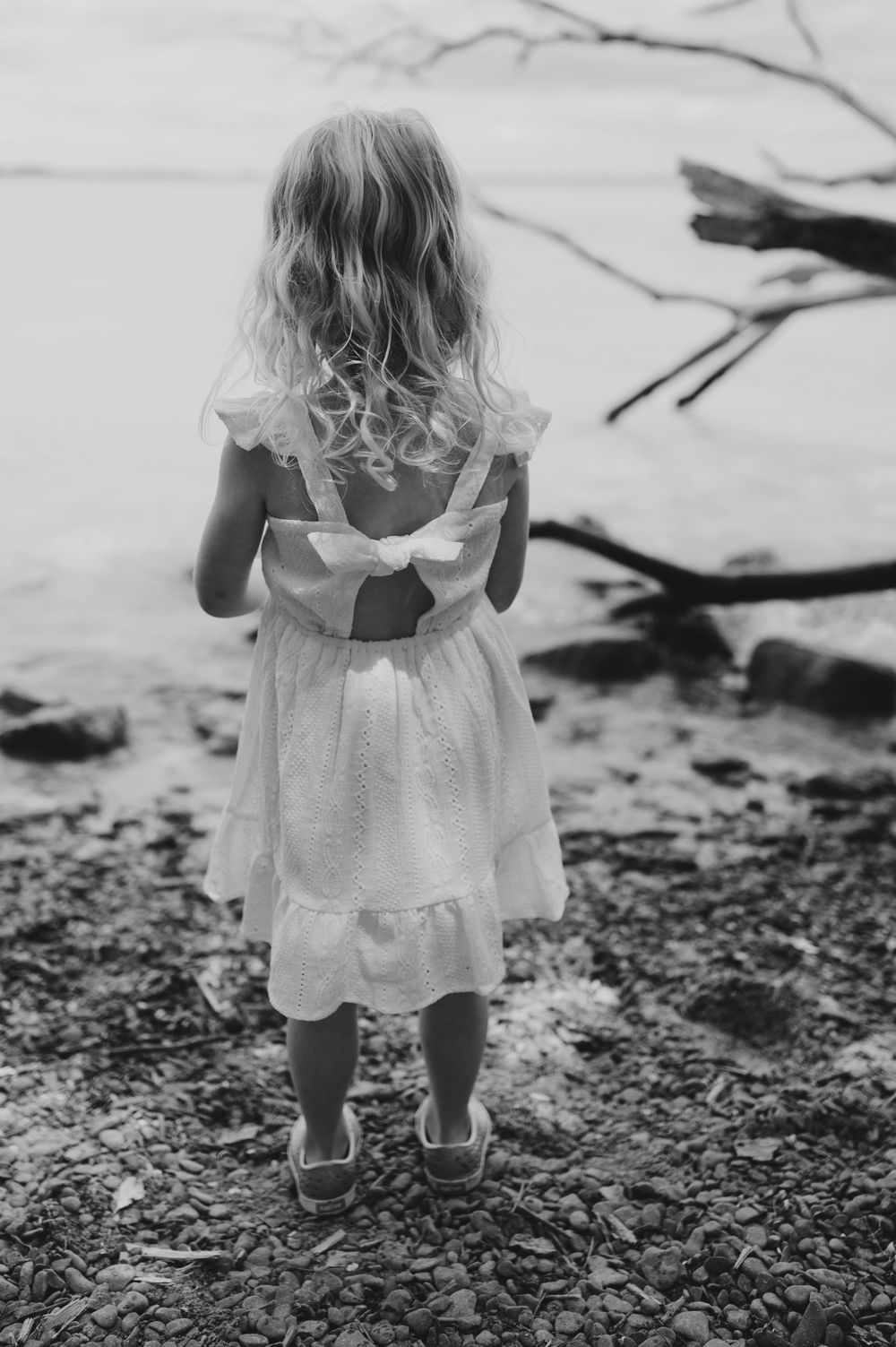 A child in a white dress stands on a rocky shore looking out at the water.