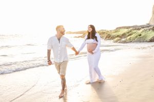 Romantic couple walking hand in hand along a sunny beach in white outfits.