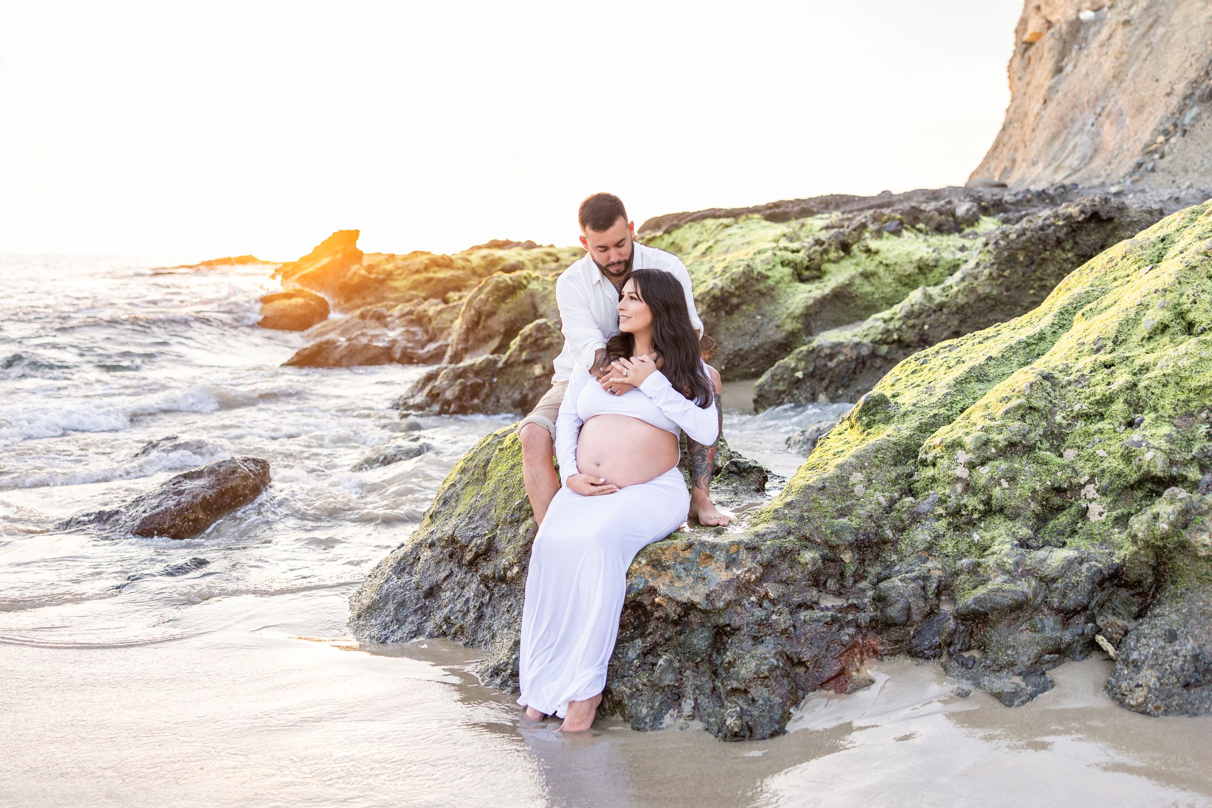 Couple in white clothing shares intimate moments on a rocky beach during golden hour.