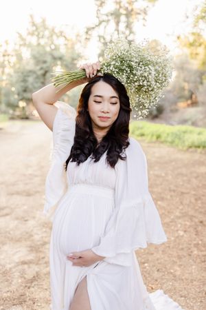 Expectant mother in white dress holding baby's breath flowers against a natural outdoor backdrop.