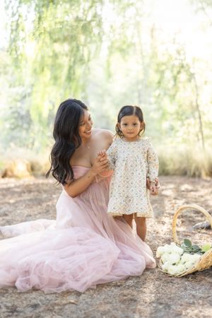 Mother and child in pink and white dresses share tender moment under willow trees.