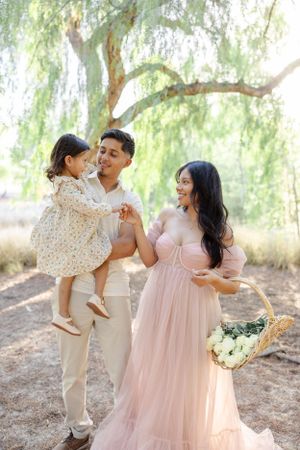 Family portrait under a weeping willow tree with woman in pink dress holding floral basket in natural outdoor setting.