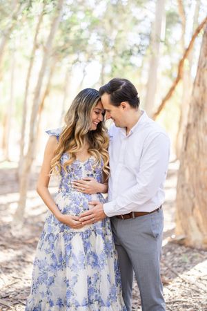 Maternity photo in forest with couple wearing blue floral dress and white shirt.