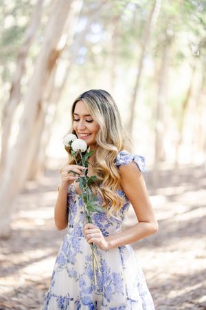 Woman in blue floral dress smelling white roses in sunlit forest setting.