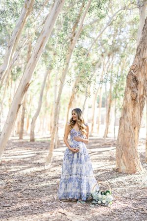 A pregnant woman in a flowing blue floral dress poses elegantly in a sunlit eucalyptus forest.