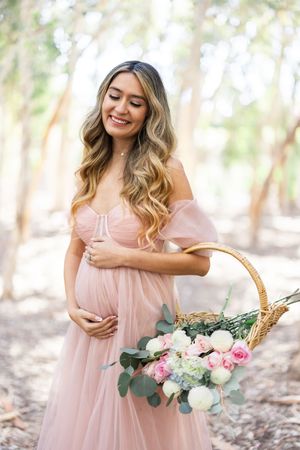 Maternity portrait with woman in pink dress holding flower basket in wooded setting.