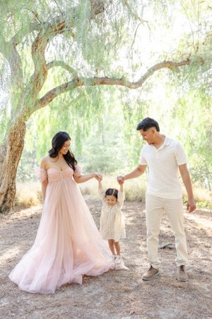 Family in white and pink outfits poses under willow trees in dreamy outdoor setting.