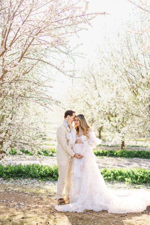 A romantic couple in white attire share tender moments under blooming spring trees in a dreamy outdoor setting.