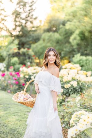 Woman in flowing white dress walks through garden of colorful roses with basket.