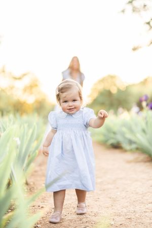 Small child in white dress toddles along garden path surrounded by green plants.