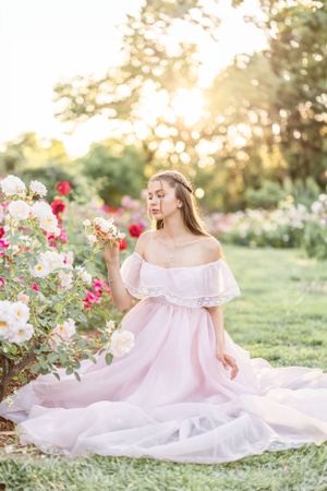 Woman in flowing pink dress sits gracefully among blooming roses in garden at sunset.
