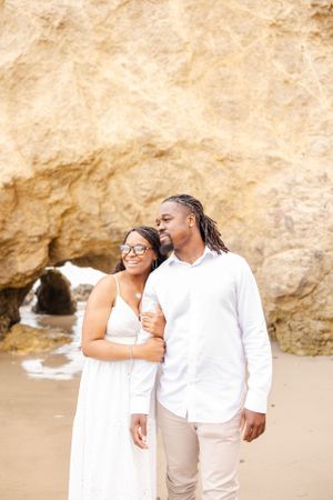 A couple in white outfits embraces on a sandy beach with rocky cliffs and ocean waves in the background.