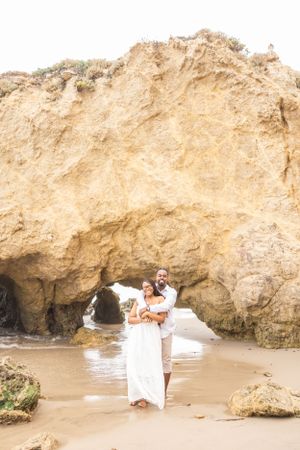 A romantic couple embraces on a sandy beach with dramatic limestone cliffs and caves in the background.