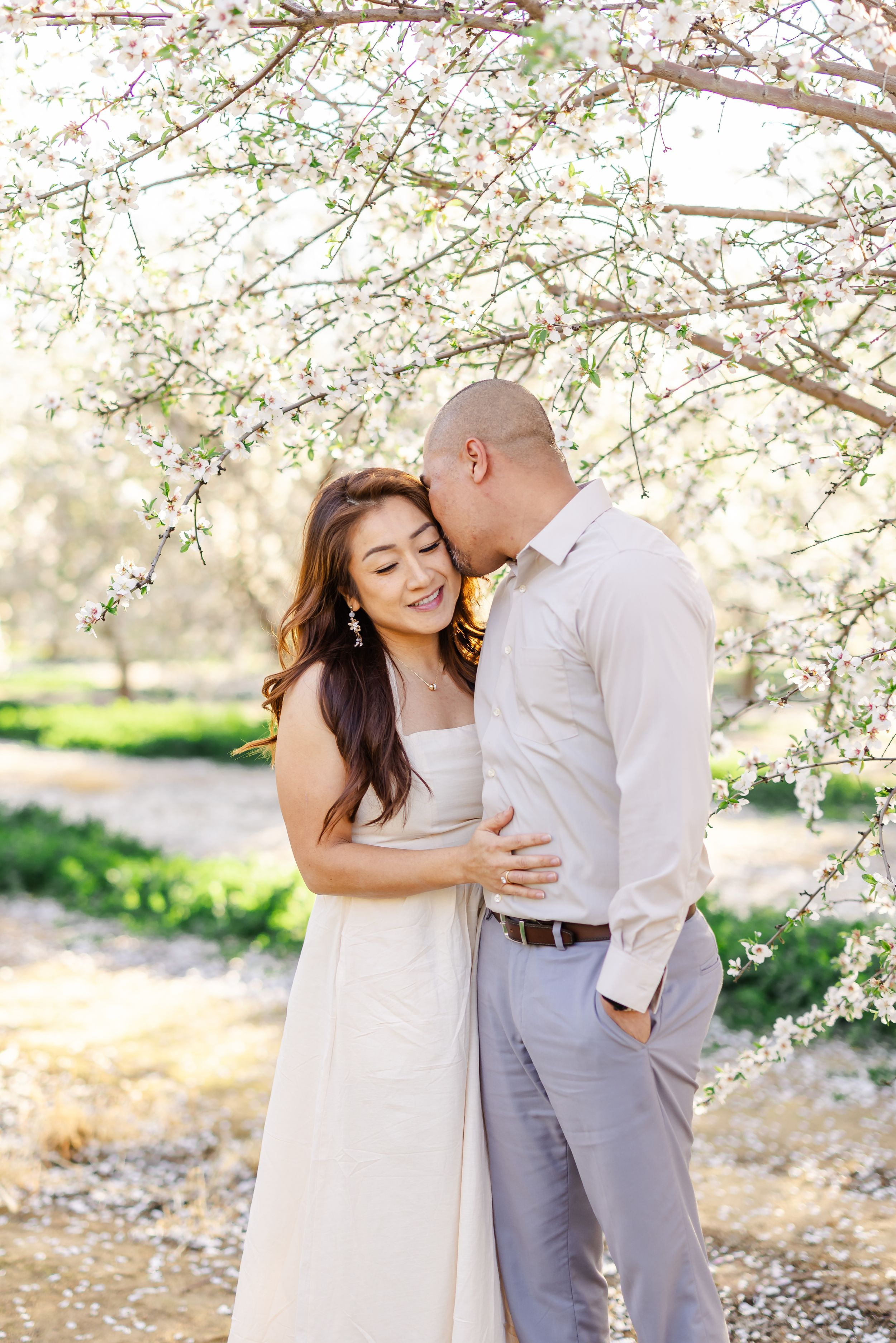 Couple embracing under white cherry blossom tree, woman in white dress smiling as man kisses her cheek outdoors.