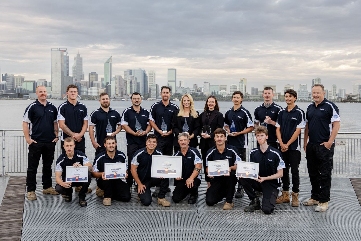 Team in matching black uniforms poses together on waterfront deck with city skyline backdrop at dusk.