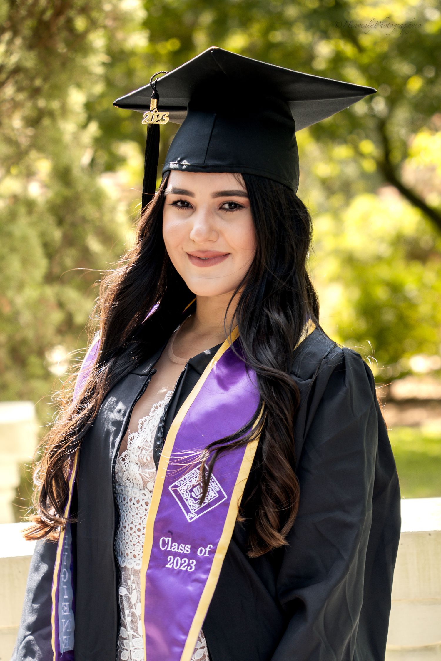 Graduate in cap and gown with purple sash poses in natural outdoor setting.