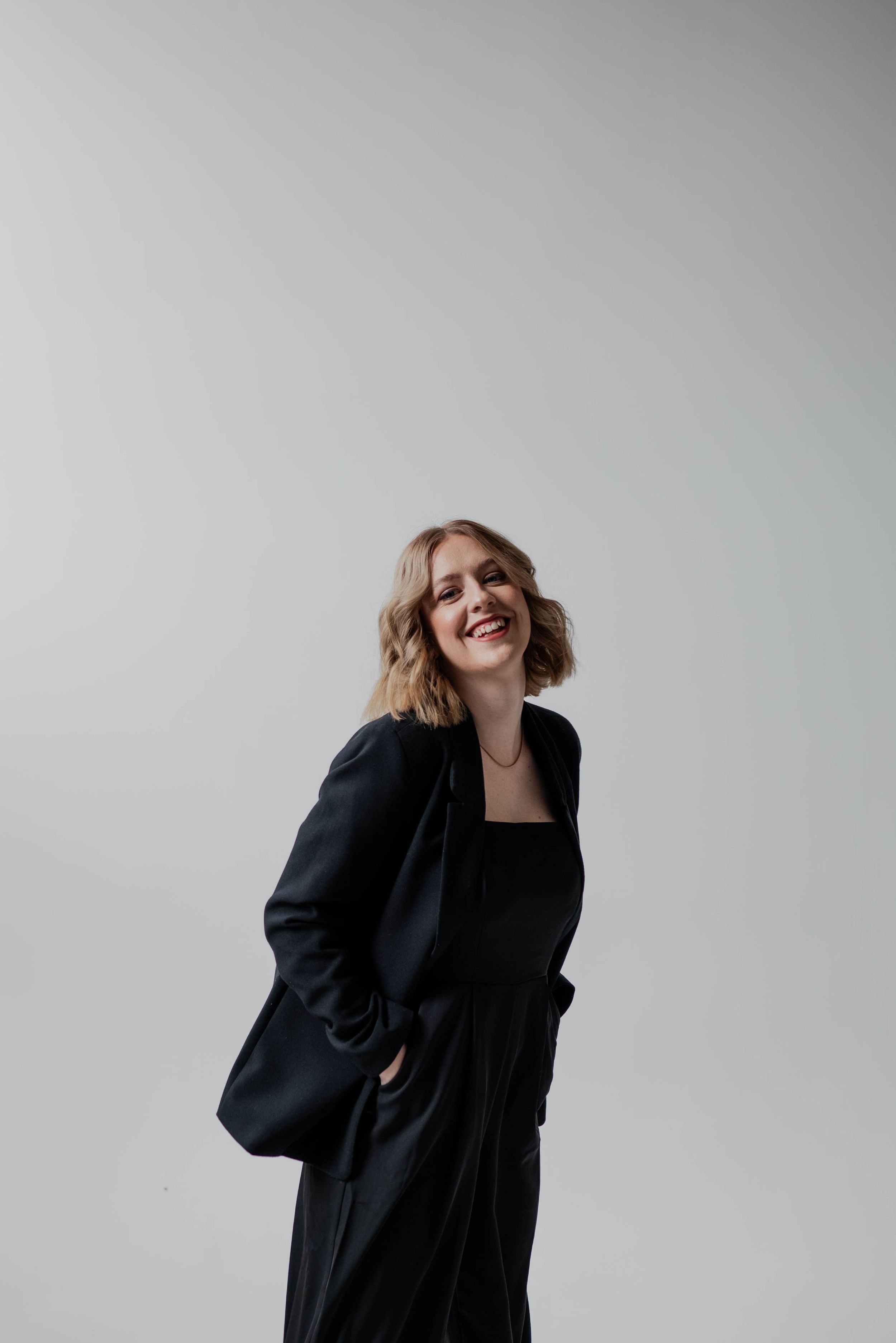 Woman in black blazer and dress smiles joyfully against white background.