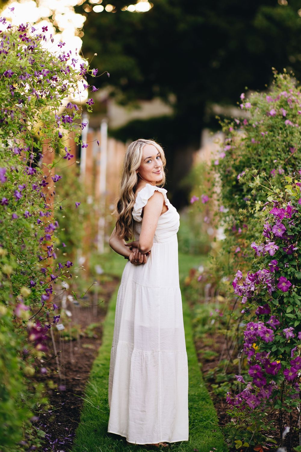 Young girl in white dress twirls and plays in a garden with blooming purple flowers and greenery.