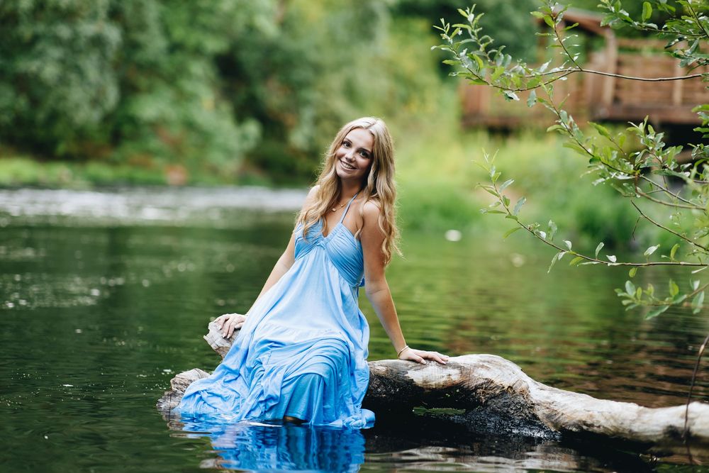 A person in a flowing blue dress poses gracefully near a tranquil river surrounded by lush greenery.