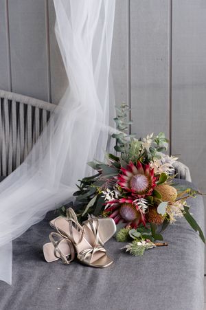 A bridal bouquet with protea flowers and eucalyptus rests beside nude strappy heels on grey fabric with flowing white veil.