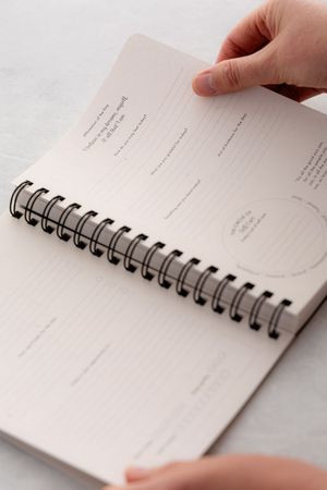 Hands hold open a spiral-bound notebook with blank pages and printed text forms on white surface.