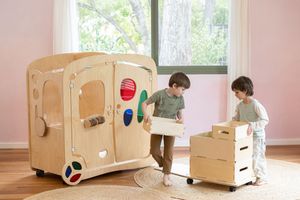 Children playing with large cardboard playhouses on wheels in a bright sunlit room with pink walls.