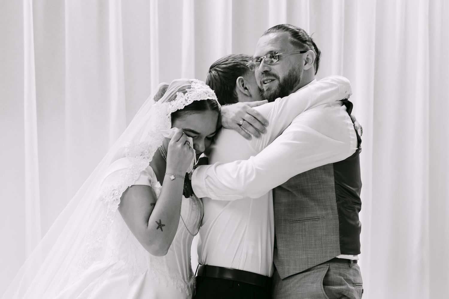 Black and white photo of emotional bride and groom sharing a group hug with friend at their wedding ceremony.