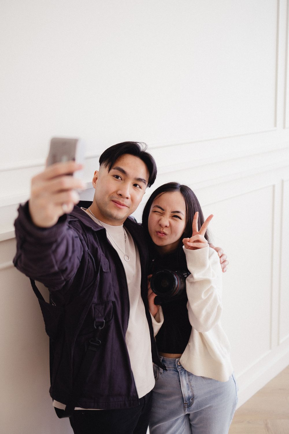 Couple taking selfies against white wall in casual outfits.