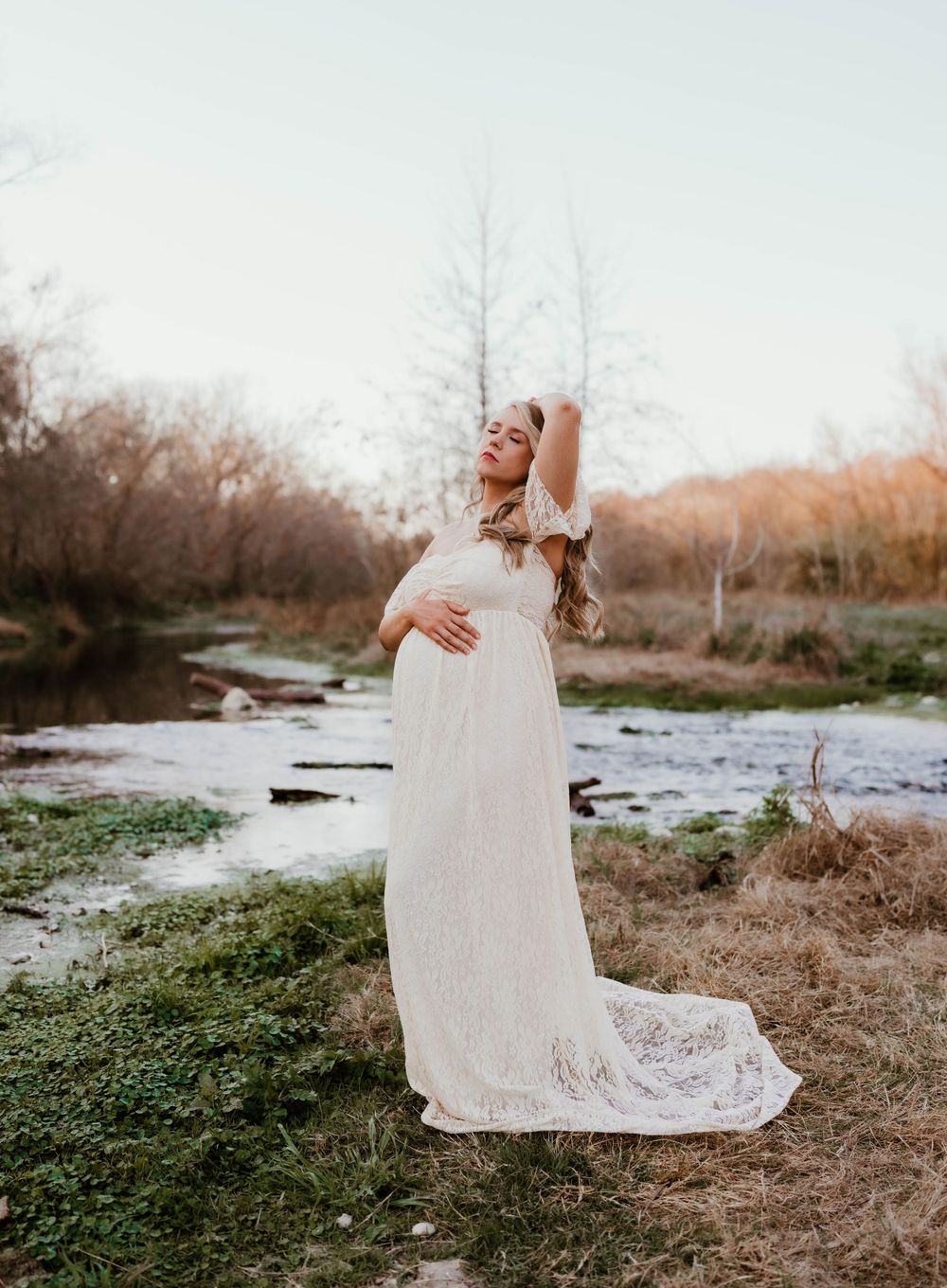 Pregnant woman in flowing white dress poses elegantly by a serene stream at dusk.