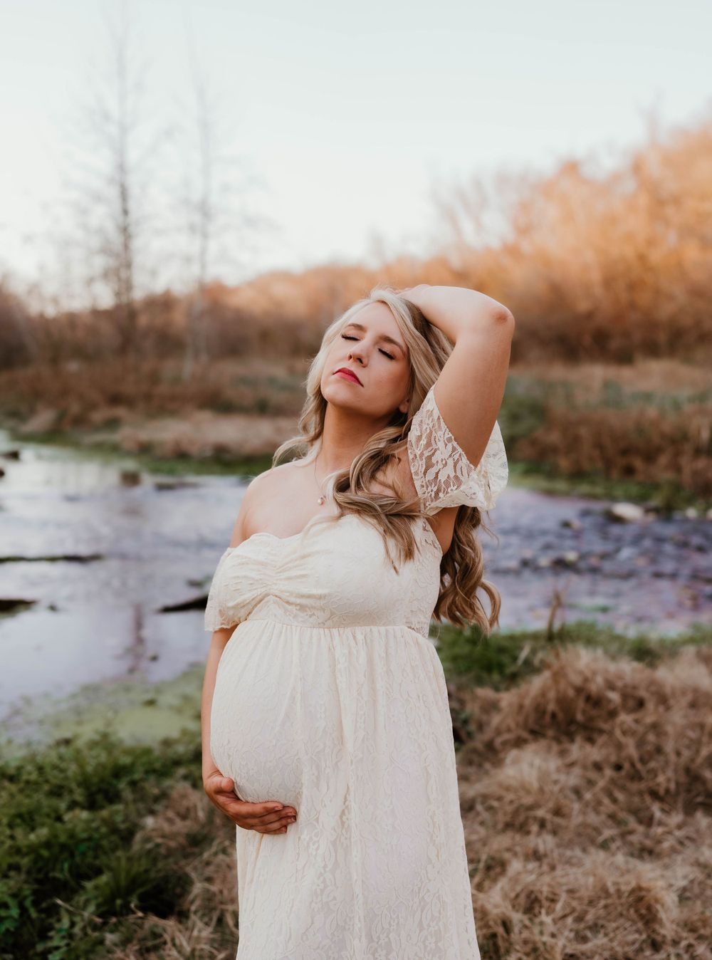 Expectant mother in white lace dress poses gracefully by a stream during sunset.