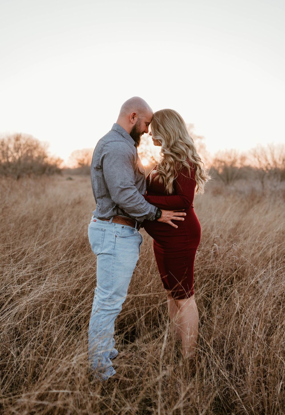 Sweet maternity moment between couple standing in tall grass during sunset.