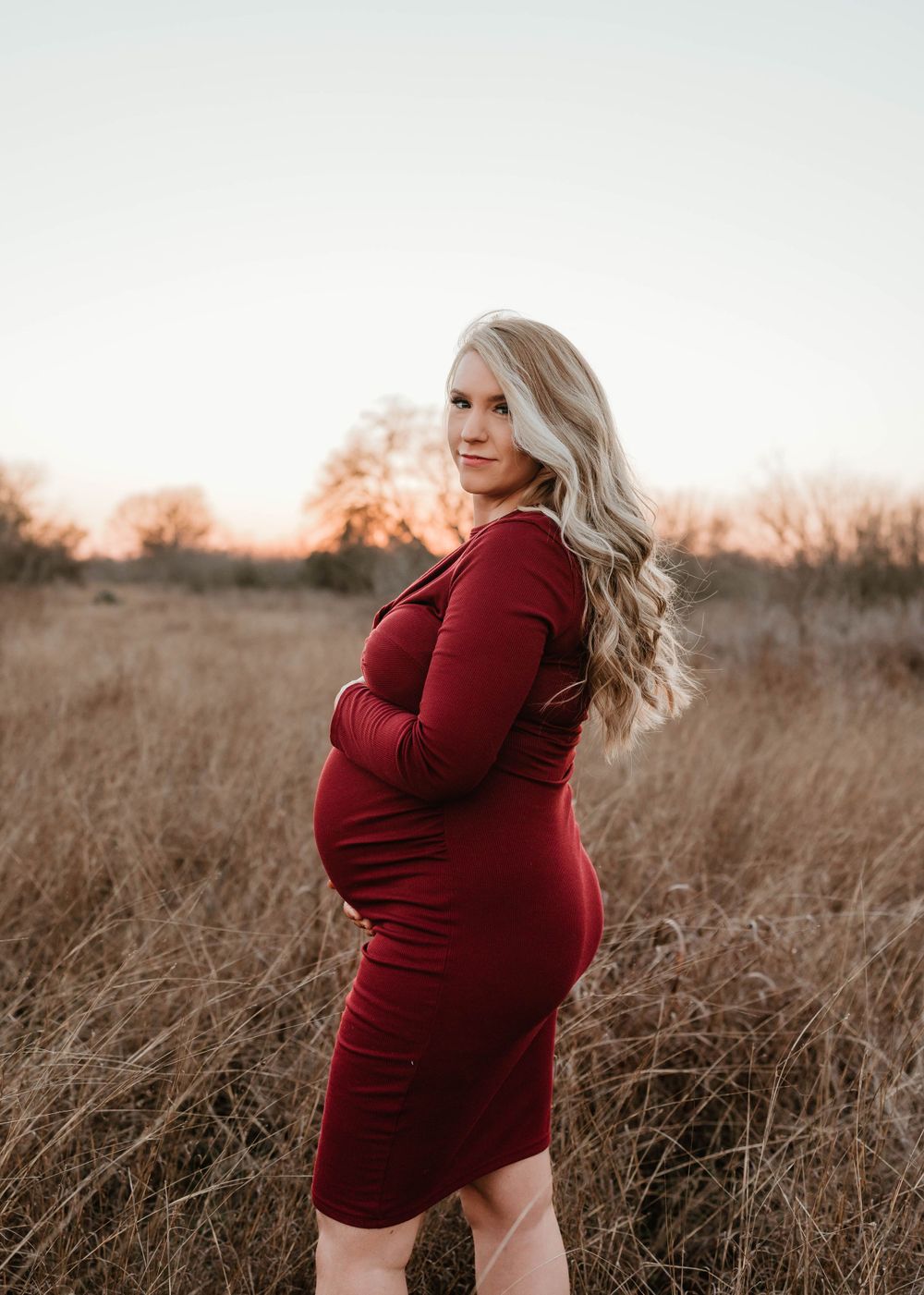 A pregnant woman in a burgundy dress poses in a field of tall grass during golden hour.