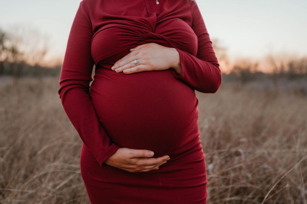 A pregnant woman in a burgundy dress cradles her baby bump while standing in a golden autumn field at sunset.