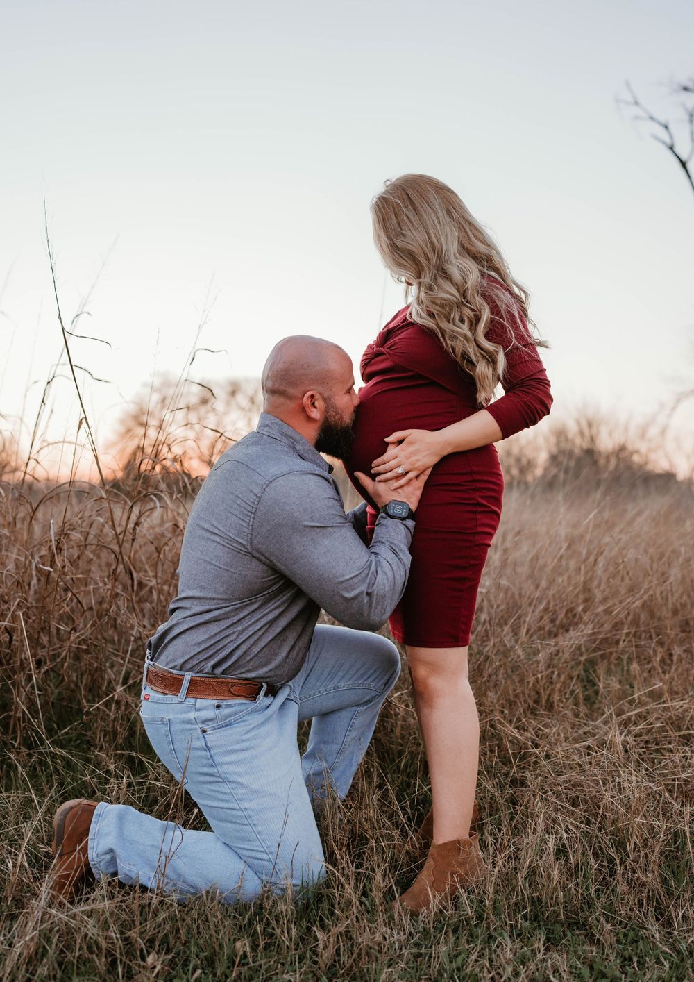 A person wearing gray kneels beside someone in a burgundy dress in a field of dried grass at sunset.