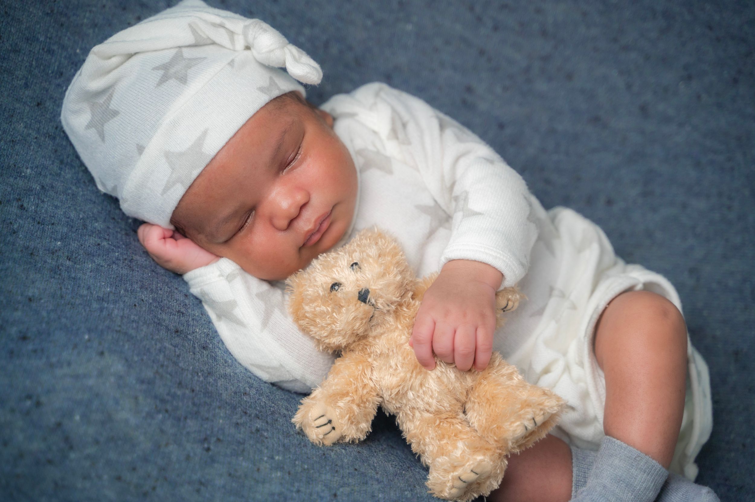 Sleeping baby in white outfit cuddles with small teddy bear on blue blanket.