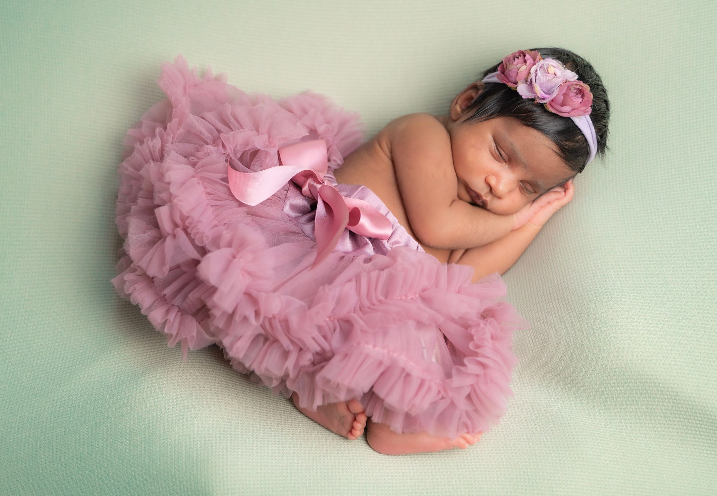 Sleeping newborn wearing pink tutu and floral headband on soft mint background.