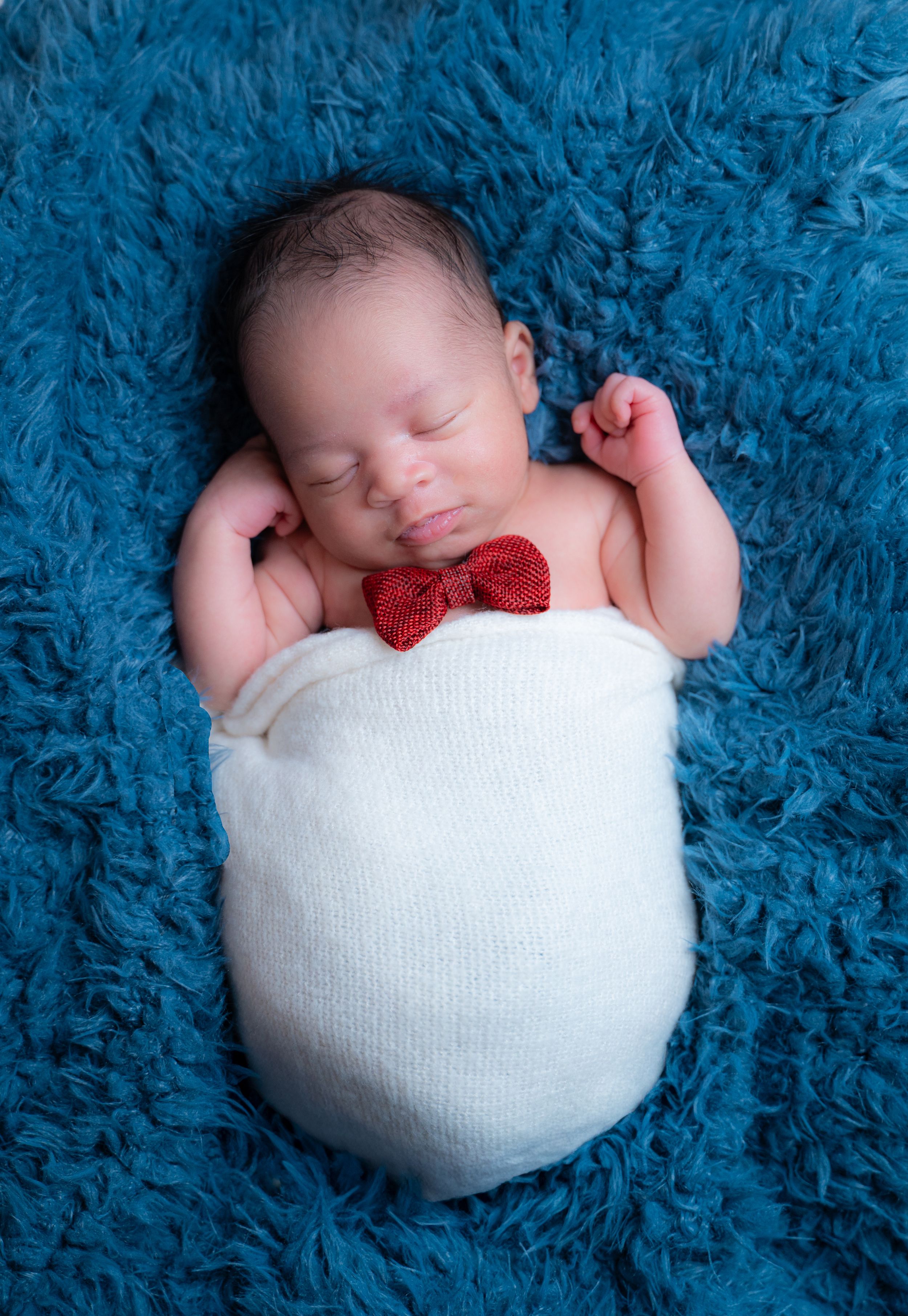 Sleeping newborn swaddled in white fabric with red bowtie lying on soft blue furry blanket in professional photo shoot setting.