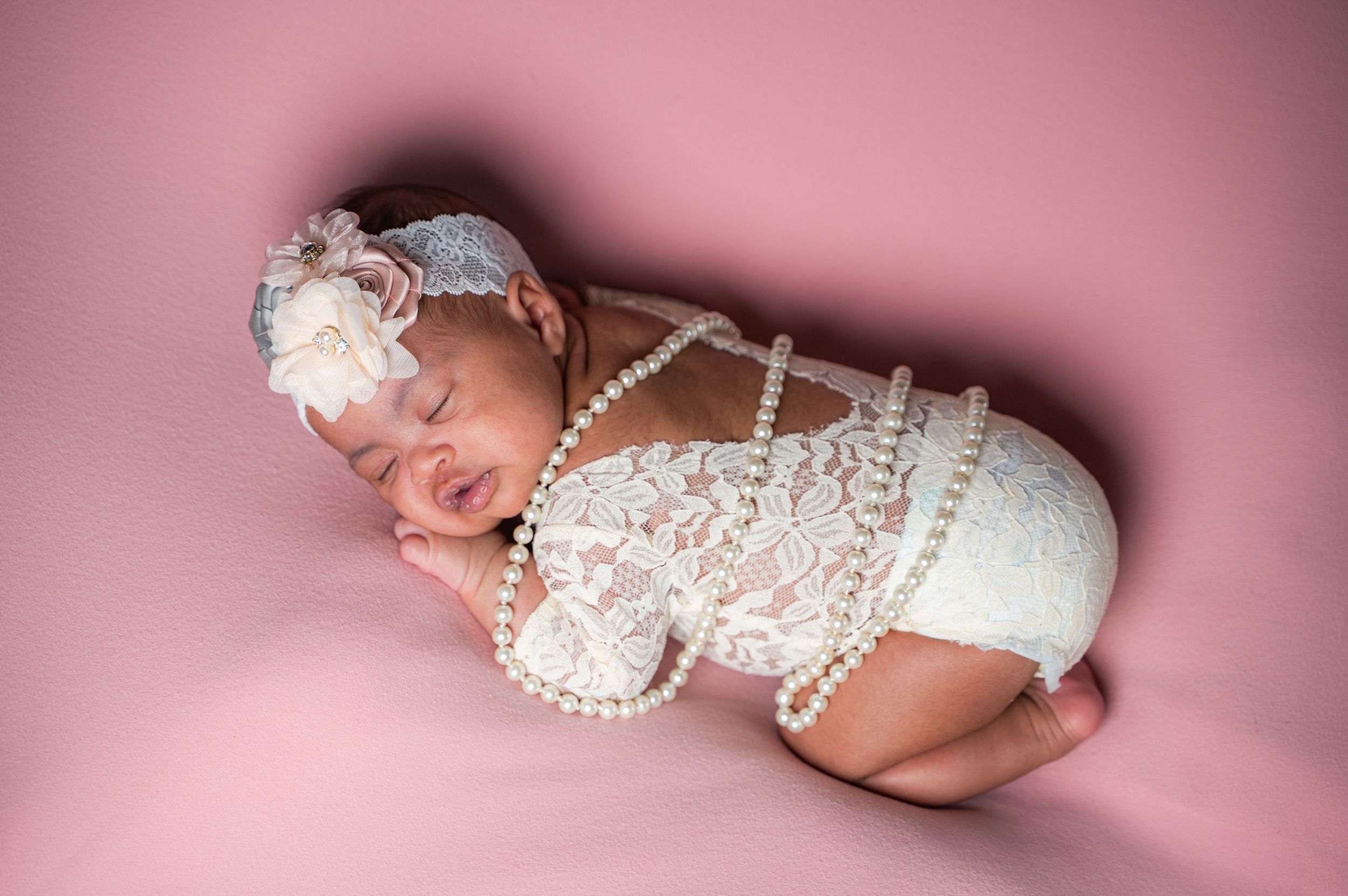 Baby in delicate white lace outfit sleeping peacefully on pink background.