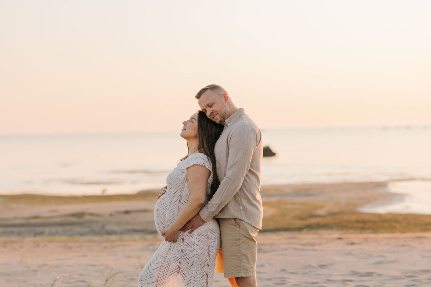 Couple embraces on beach at sunset with expecting mother in flowy dress.