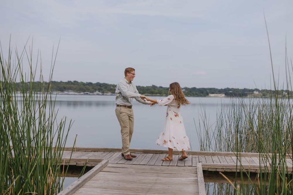 Couple holding hands and spinning on a wooden dock with a serene lake and tall grass in background.