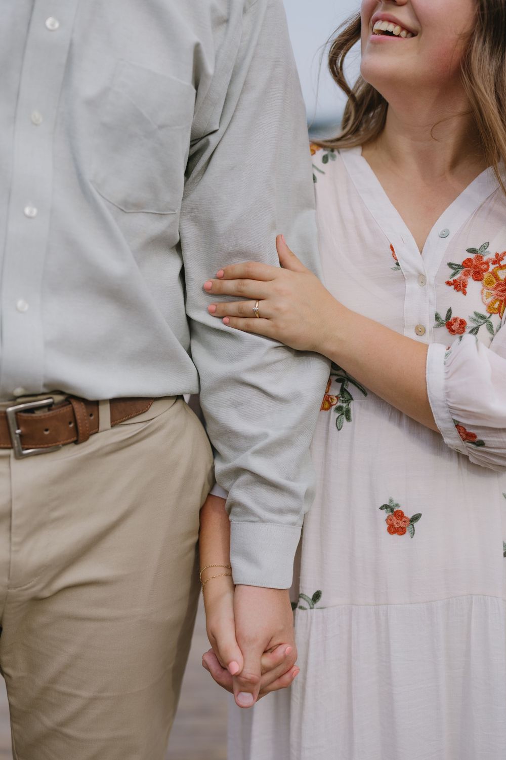 Close-up of couple holding hands, woman in white floral dress with engagement ring, man in khakis.